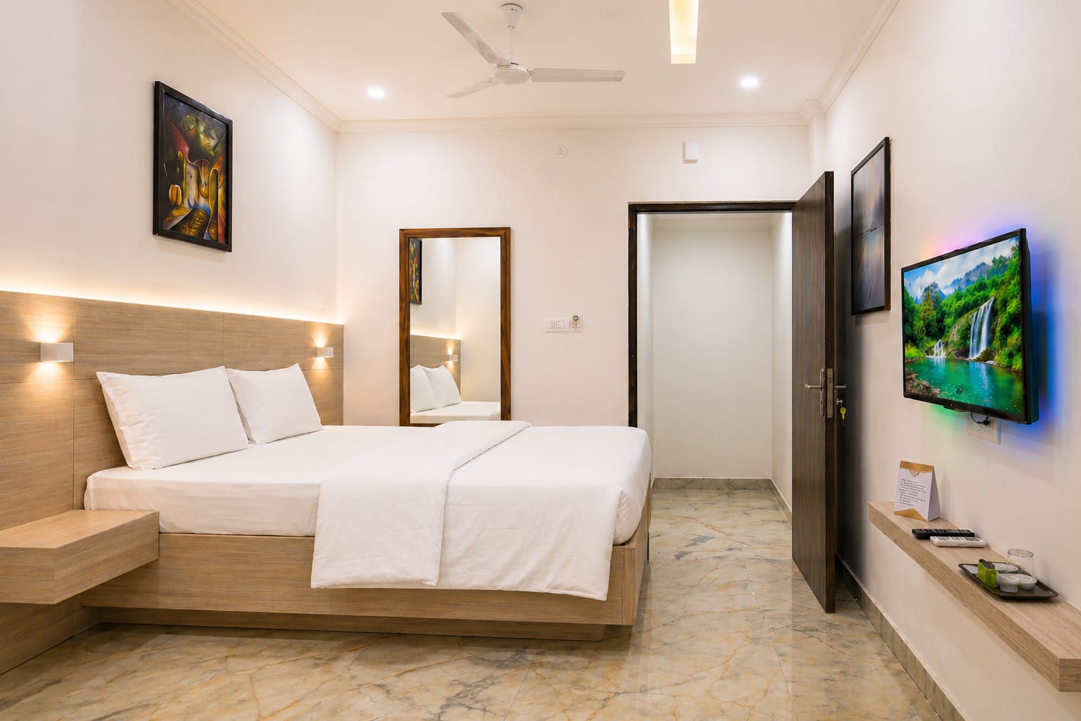 Rooms A modern, clean hotel room at Sri Aditya Inn featuring a queen-sized bed with white linens, light wood headboard with warm accent lighting, a wall-mounted TV with colorful LED backglow, and polished marble floors.